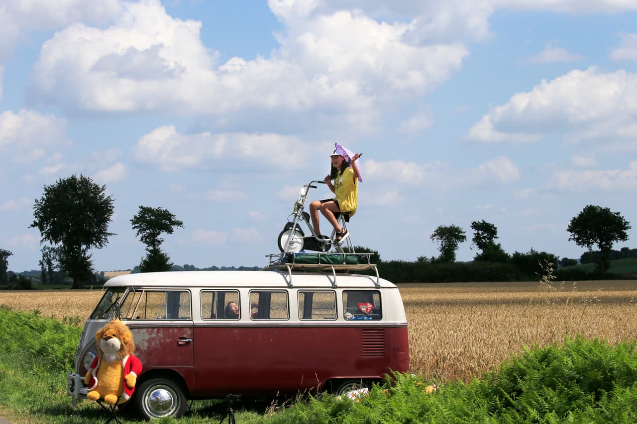 Encima de una camioneta de pasajeros aparece, montada en una bicicleta, una niña, quien espera por el paso de los ciclistas al lado de la carrertera en el occidente de Francia.