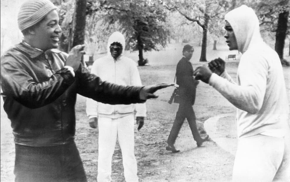 El 12 de mayo de 1966 en Hyde Park, Londres, Muhammad Ali pretende algunos rounds de entrenamiento con su gran amigo el corredor Jim Brown de Cleveland Browns. Al fondo está Jimmy Ellis, entonces sparring de Ali, luego uno de sus rivales.