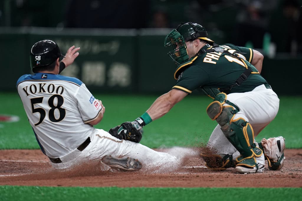 Toshitake Yokoo de los Hokkaido Nippon-Ham Fighters es puesto out en el plato por el receptor Josh Phegley de los Oakland Athletics en una jugada espectacular donde la tierra se levanta.