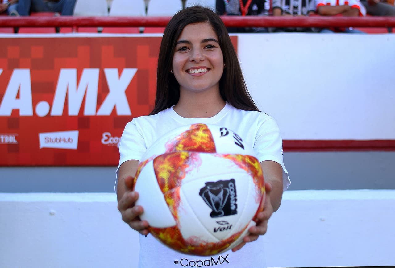 La chica elegida para el protocolo de la entrega del esférico muestra sonriente la herramienta de trabajo para esta instancia de la competencia copera.