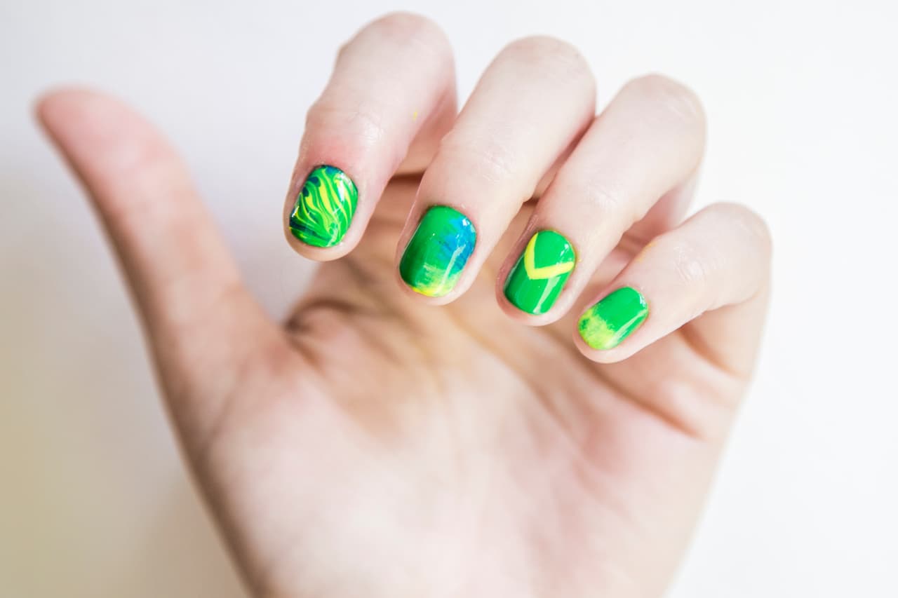 Brasil: Aplica 2 capas de tono verde en las uñas. Dibuja una línea diagonal verde, azul y amarillo en la uña del dedo índice y sin esperar a que seque mezcla los tonos con un punzón formando ochos. Con la mitad de una brocha mini abanico con esmalte amarillo y resto en azul, barre de un lado al centro. En el anular haz una V con amarillo. Aplica en un quesito una cantidad pequeña de esmalte amarillo y da toques en la punta de la uña del meñique.