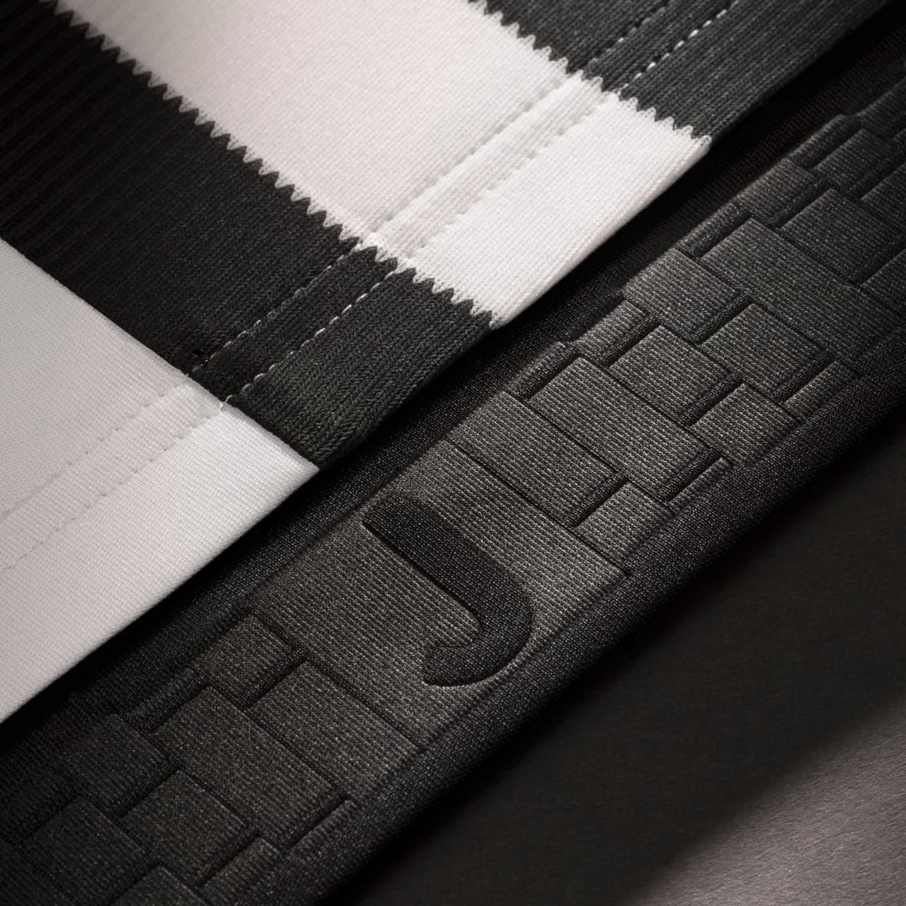 El cintillo horizontal negro en la parte inferior de la camiseta muestra un diseño gráfico destacando la letra J, simbólica del club, y detalles que evocan el emblemático exterior del estadio de la Juventus.