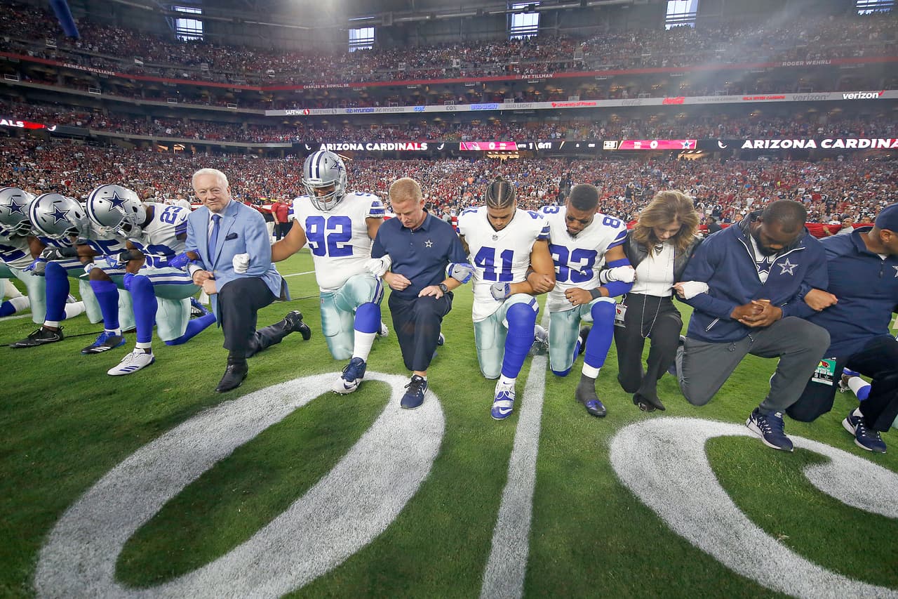 Dallas Cowboys owner Jerry Jones (in jacket) and head coach Jason Garrett (blue shirt) kneel with their team in a show of solidarity before the national anthem during a 2017 NFL week 3 regular season game against the Arizona Cardinals, Monday, Sept. 25, 2017 in Tempe, Ariz. (James D. Smith via AP)