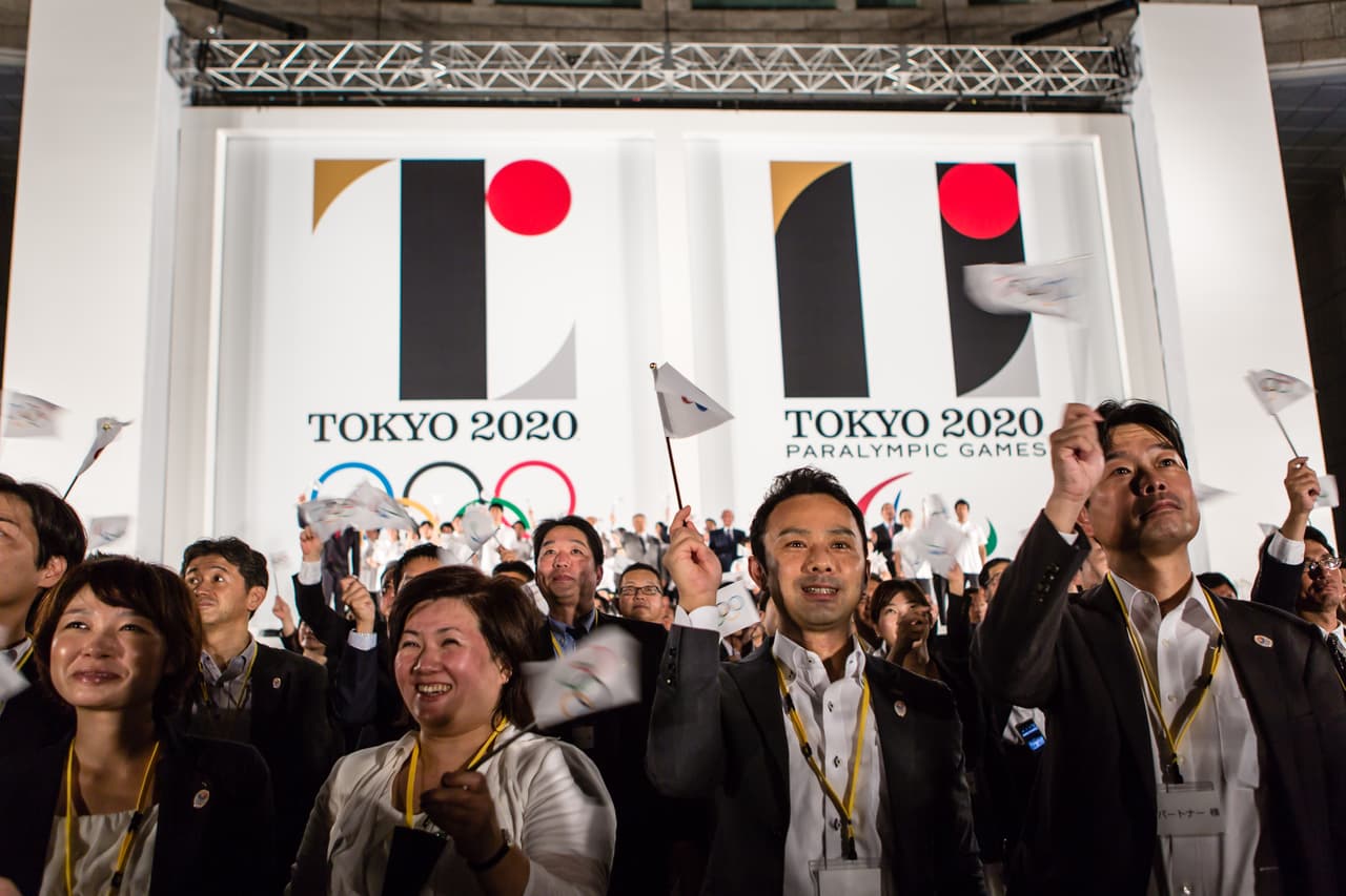 TOKYO, JAPAN - JULY 24: People wave flags as the new Olympic emblems are unveiled during a ceremony for the Tokyo 2020 Olympic and Paralympic Games at the Tokyo Metropolitan Government Plaza on July 24, 2015 in Tokyo, Japan. The emblem for Tokyo 2020 symbolizes the power of unity. The black color of the center column represents diversity, which is a combination of all colors. The circular shape represents an inclusive world where everyone accepts each other. The red part of the circle represents strength of every beating heart. With all these elements combined, creates the emblems for the Olympic and Paralympic Games. The Tokyo 2020 Olympic Emblem is inspired by the "T" in Tokyo Tomorrow Team and the Tokyo 2020 Paralympic emblem is inspired by the universal sign of equality. (Photo by Christopher Jue/Getty Images)