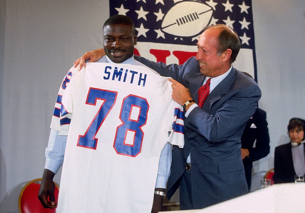 1985 BRUCE SMITH | DT Virginia Tech - 
<b>Buffalo Bills</b>
<br>
<br>El comisionado de la NFL, Pete Rozelle, sostiene el nuevo jersey para el recién elegido por Buffalo, Bruce Smith (78) ala defensiva, primera selección de los Bills y la selección número uno global de la NFL celebrado el 30 de abril de 1985 en Nueva York.