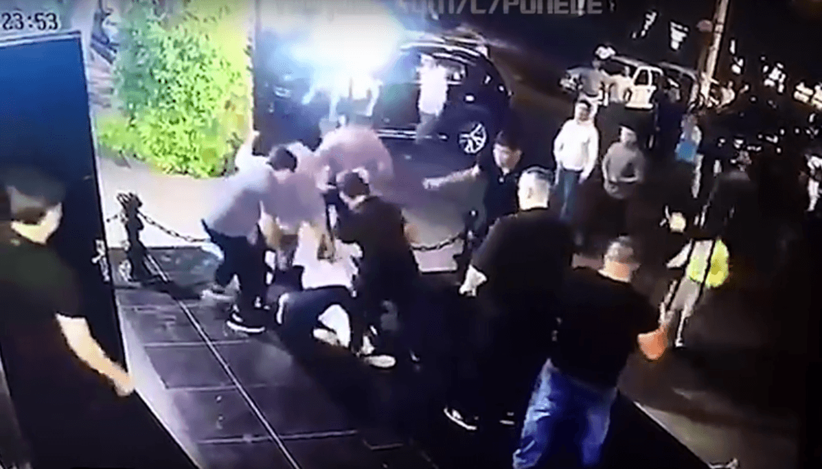 En mayoría numérica, uno de los guardias agarra con una llave de piernas a Dzhamshid Kenzháyev, vestido de blanco. Otro de los que está de pie patea al luchador.